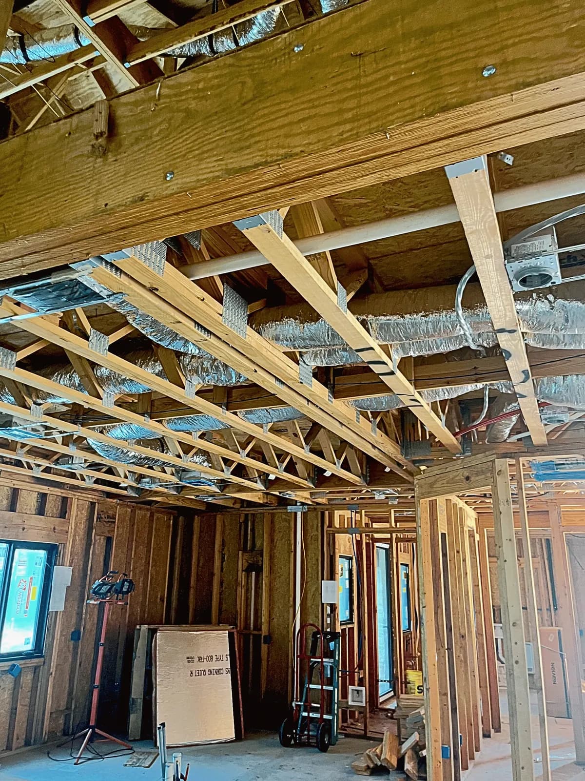 Insulated flexible duct branches threaded through the ceiling framing of a house under construction, with recessed light housings and wiring roughed in above bare stud walls.